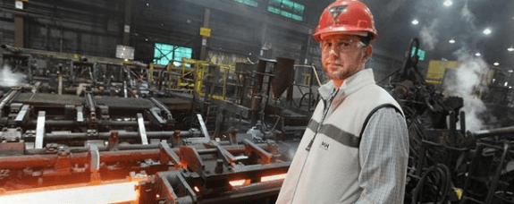 A man with a hard hat in a factory. Behind him is red hot steel